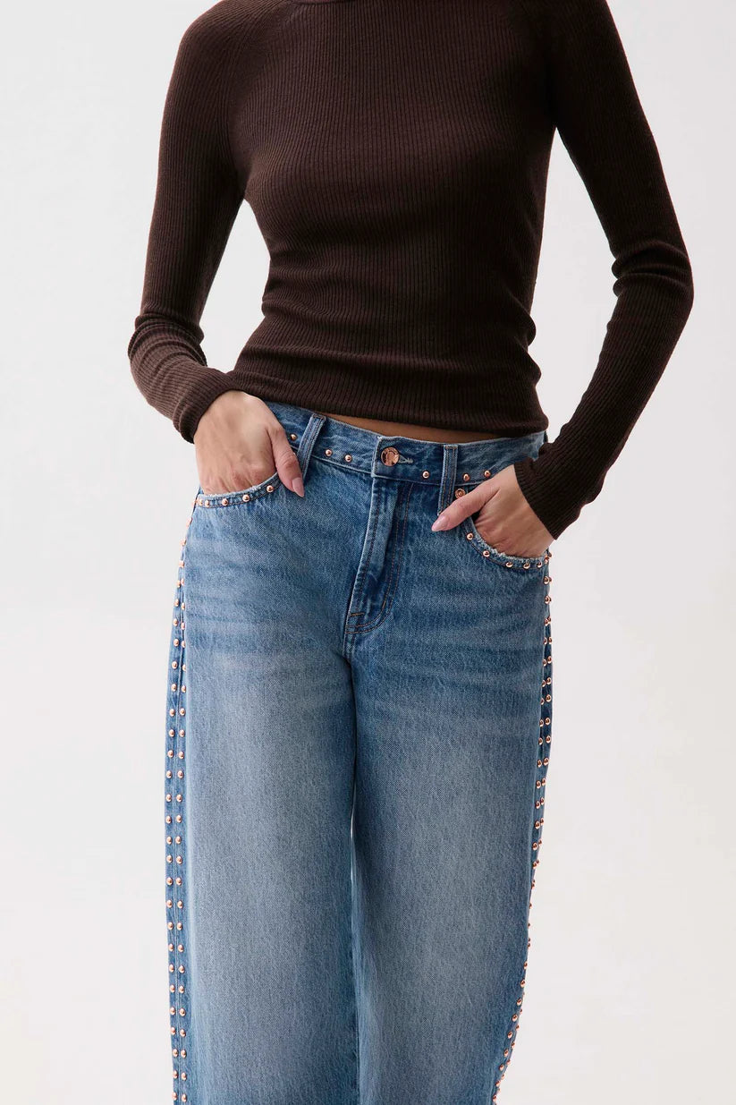 A model wearing a dark brown ribbed long-sleeve top and light blue jeans with gold-tone stud detailing along the outer seams of the legs, standing against a plain white background.