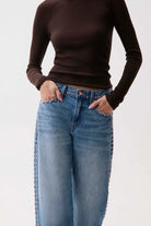 A model wearing a dark brown ribbed long-sleeve top and light blue jeans with gold-tone stud detailing along the outer seams of the legs, standing against a plain white background.