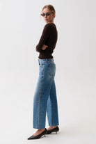 A woman wearing blue wide-leg jeans with silver studs down the side, a dark brown long-sleeve top, and black pointed-toe heels, photographed against a plain light gray studio background.