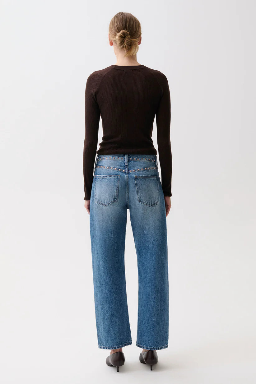 A woman viewed from behind wearing a dark brown long-sleeve top, blue wide-leg jeans with silver studs on the waistband, and black heels, set against a plain white studio background.