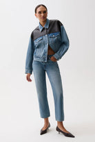 A model wearing a denim jacket with a black leather yoke and matching blue denim jeans, styled with black pointed-toe heels against a plain white studio background.