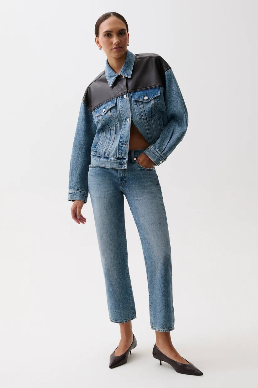 A model wearing a denim jacket with a black leather yoke and matching blue denim jeans, styled with black pointed-toe heels against a plain white studio background.