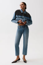 A model wearing a cropped denim jacket with a color-blocked design in medium and dark blue, paired with high-waisted blue jeans and black pointed-toe heels, against a plain white studio background.