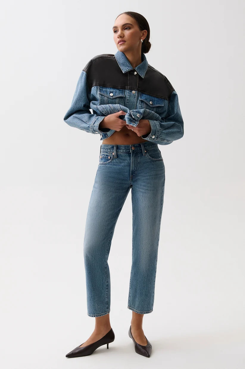 A model wearing a cropped denim jacket with a color-blocked design in medium and dark blue, paired with high-waisted blue jeans and black pointed-toe heels, against a plain white studio background.