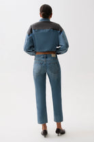 A model viewed from the back wearing a cropped denim jacket with contrasting black shoulder panels and blue jeans, paired with black heels, against a plain white studio background.