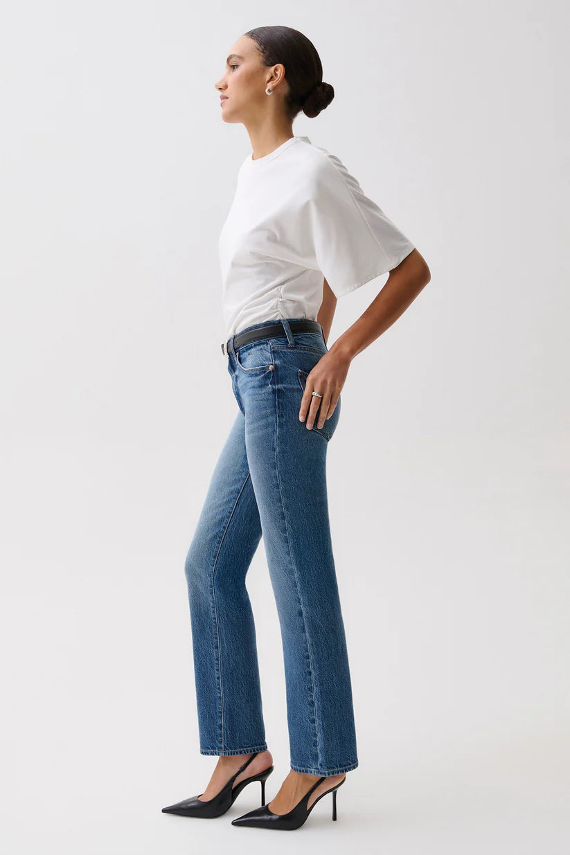 A woman in side profile wearing blue high-waisted flare jeans, a white short-sleeve top, and black heels against a light gray studio background.
