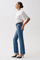 A woman in side profile wearing blue high-waisted flare jeans, a white short-sleeve top, and black heels against a light gray studio background.