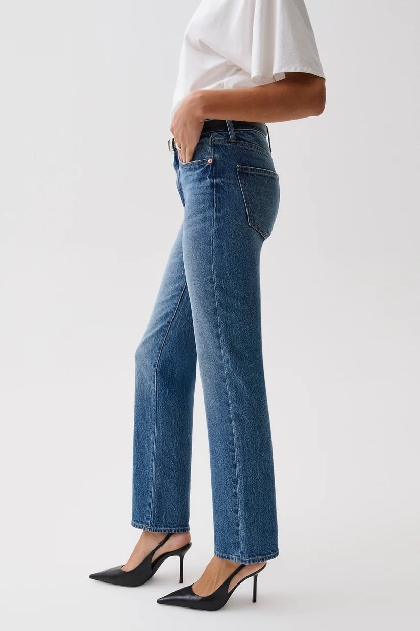 A side profile view of a model wearing medium-wash blue straight-leg jeans, a white top, and black pointed-toe mule heels against a light gray studio background.