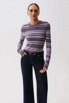 A woman modeling a long-sleeve crewneck knit top with horizontal stripes in purple, pink, and gray, paired with dark blue jeans and a black belt, against a plain light gray background.