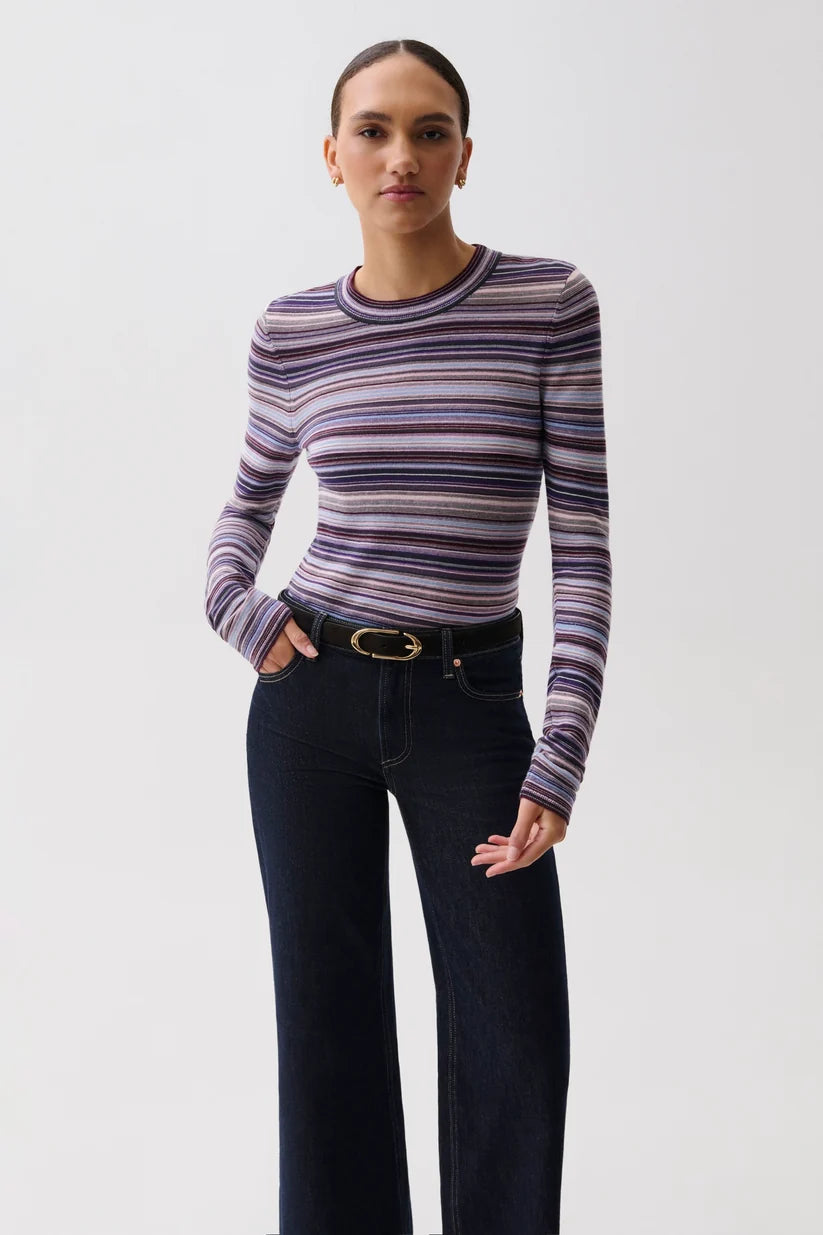 A woman modeling a long-sleeve crewneck knit top with horizontal stripes in purple, pink, and gray, paired with dark blue jeans and a black belt, against a plain light gray background.