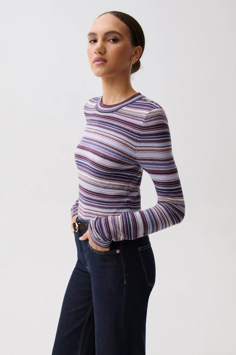 A woman modeling a long-sleeve crewneck top with horizontal purple, blue, and white stripes, paired with dark blue jeans, against a plain light gray background.
