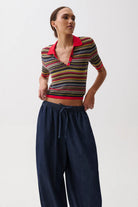 A woman modeling a short-sleeve knit polo-style top with horizontal multicolor stripes and a bright red collar, paired with dark navy drawstring trousers, against a plain light gray studio background.