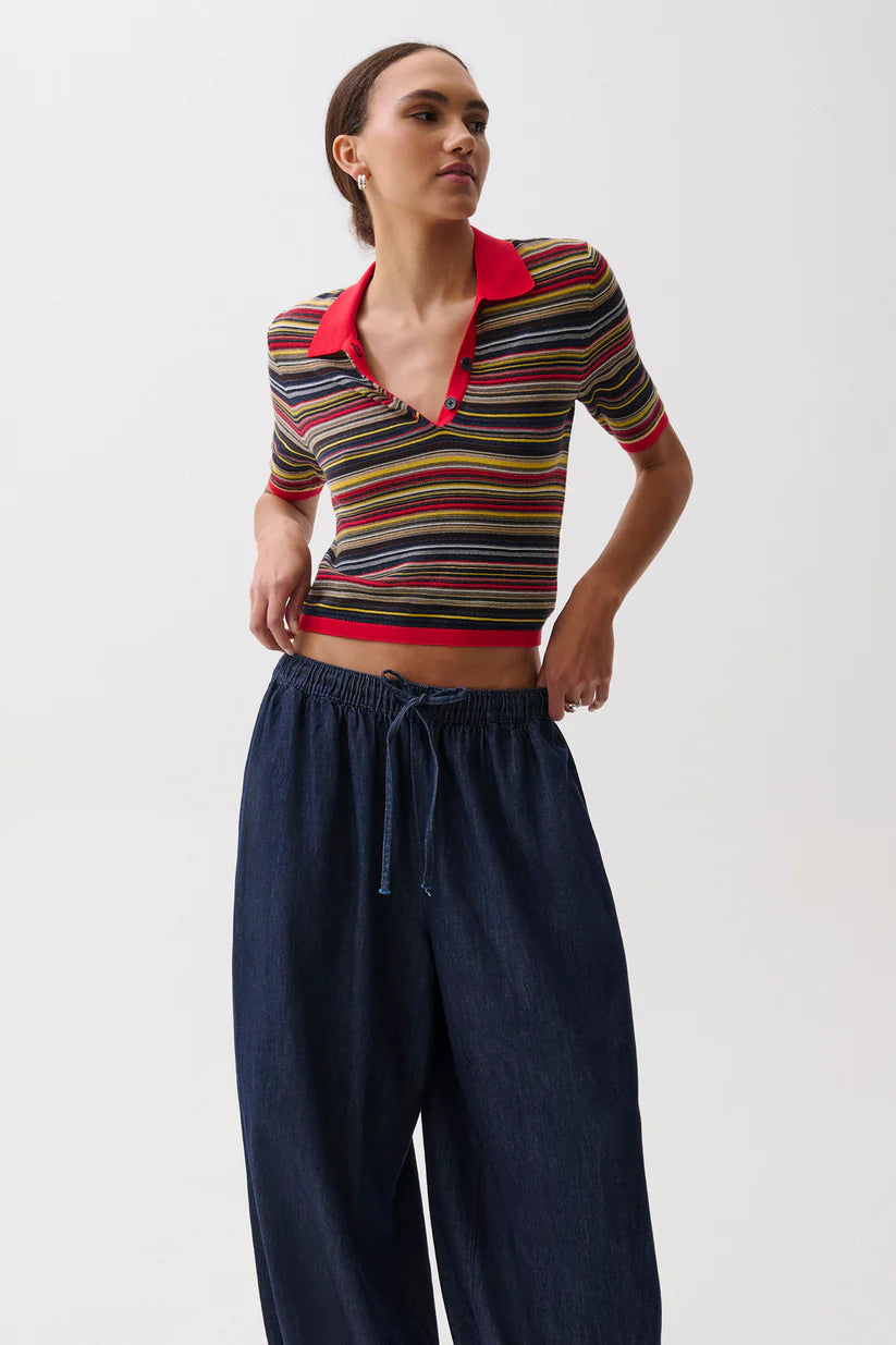 A woman modeling a short-sleeve knit polo-style top with horizontal multicolor stripes and a bright red collar, paired with dark navy drawstring trousers, against a plain light gray studio background.