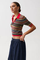 A woman modeling a short-sleeve cropped knit polo shirt with horizontal multicolor stripes and a red collar, paired with dark blue wide-leg pants, against a light gray studio background.