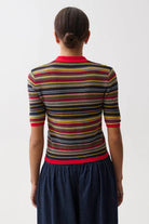 A woman shown from the back wearing a short-sleeve knit top with horizontal stripes in red, yellow, green, black, and beige, paired with a dark blue skirt, against a plain light gray background.