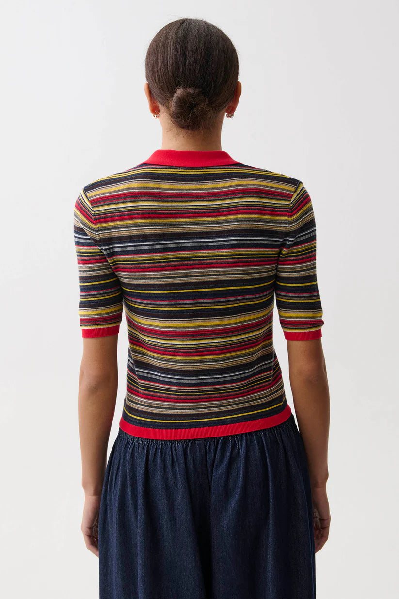 A woman shown from the back wearing a short-sleeve knit top with horizontal stripes in red, yellow, green, black, and beige, paired with a dark blue skirt, against a plain light gray background.