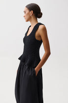 A woman in profile wearing a black sleeveless textured top with a peplum waist and black wide-leg pants, photographed against a light gray studio background.