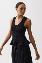 A woman modeling a black textured sleeveless top with a peplum waist, paired with a black skirt, against a plain light gray studio background.