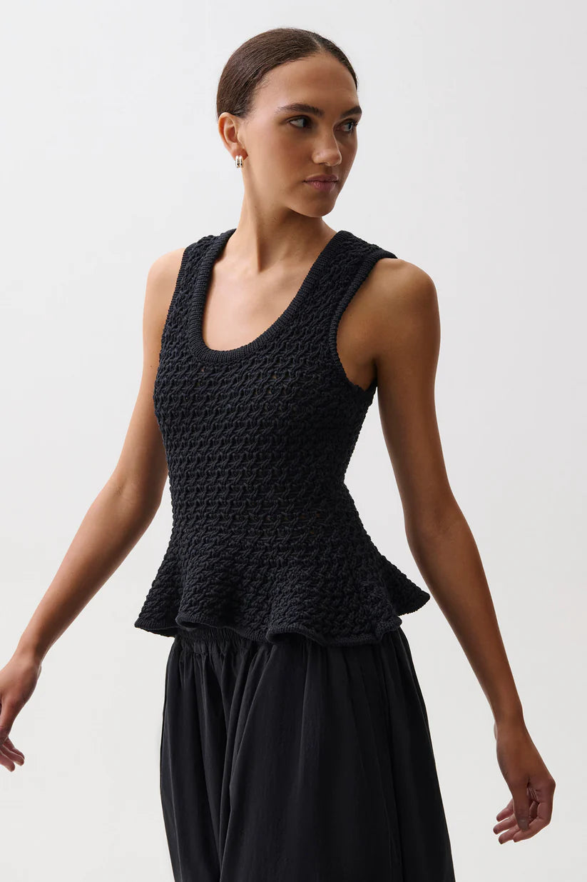 A woman modeling a black textured sleeveless top with a peplum waist, paired with a black skirt, against a plain light gray studio background.