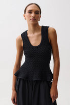 A woman modeling a black sleeveless knit top with a scoop neckline and a peplum waist, paired with a black skirt, against a plain light gray studio background.