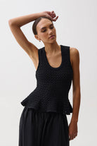 A woman modeling a black textured knit sleeveless top with a peplum waist, paired with black wide-leg trousers, against a plain light gray studio background.