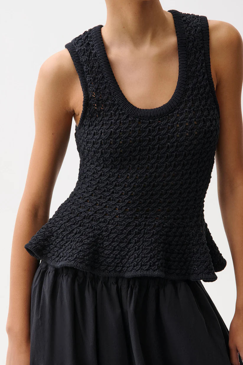 A close-up of a model wearing a black sleeveless knit top with a textured, raised pattern and a peplum hem, paired with a black skirt against a light gray background.