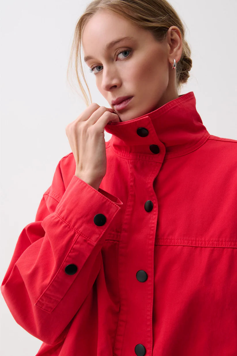 A close-up of a woman wearing a bright red high-collar jacket with black snap buttons, set against a plain light gray studio background.