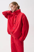 A model wearing a bright red oversized utility jumpsuit with a high collar and button front, posed against a clean light gray studio background.