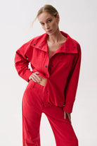 A woman modeling a bright red matching two-piece outfit consisting of a cropped button-front jacket with a wide collar and matching wide-leg pants, photographed against a clean light gray studio background.