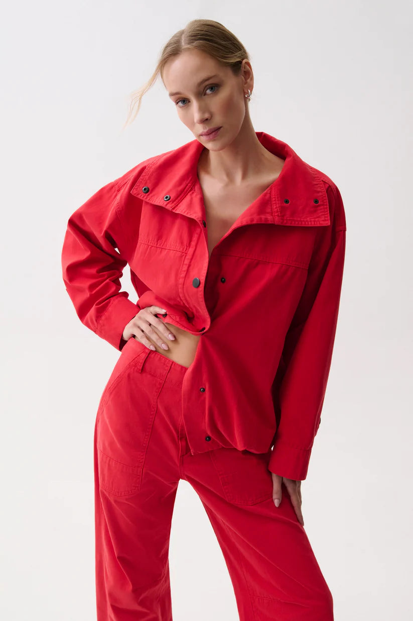 A woman modeling a bright red matching two-piece outfit consisting of a cropped button-front jacket with a wide collar and matching wide-leg pants, photographed against a clean light gray studio background.