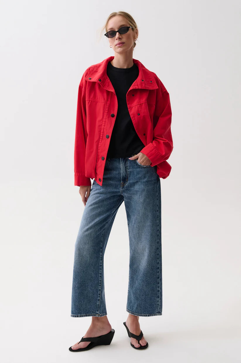 A woman modeling a bright red cropped button-up jacket over a black top, paired with wide-leg blue jeans and black wedge sandals, against a plain light gray studio background.
