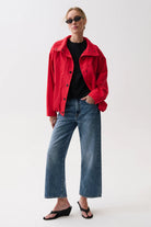 A woman modeling a bright red cropped button-up jacket over a black top, paired with wide-leg blue jeans and black wedge sandals, against a plain light gray studio background.
