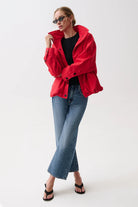 A woman modeling a bright red oversized jacket over a black top, paired with blue jeans and black sandals, against a plain white studio background.