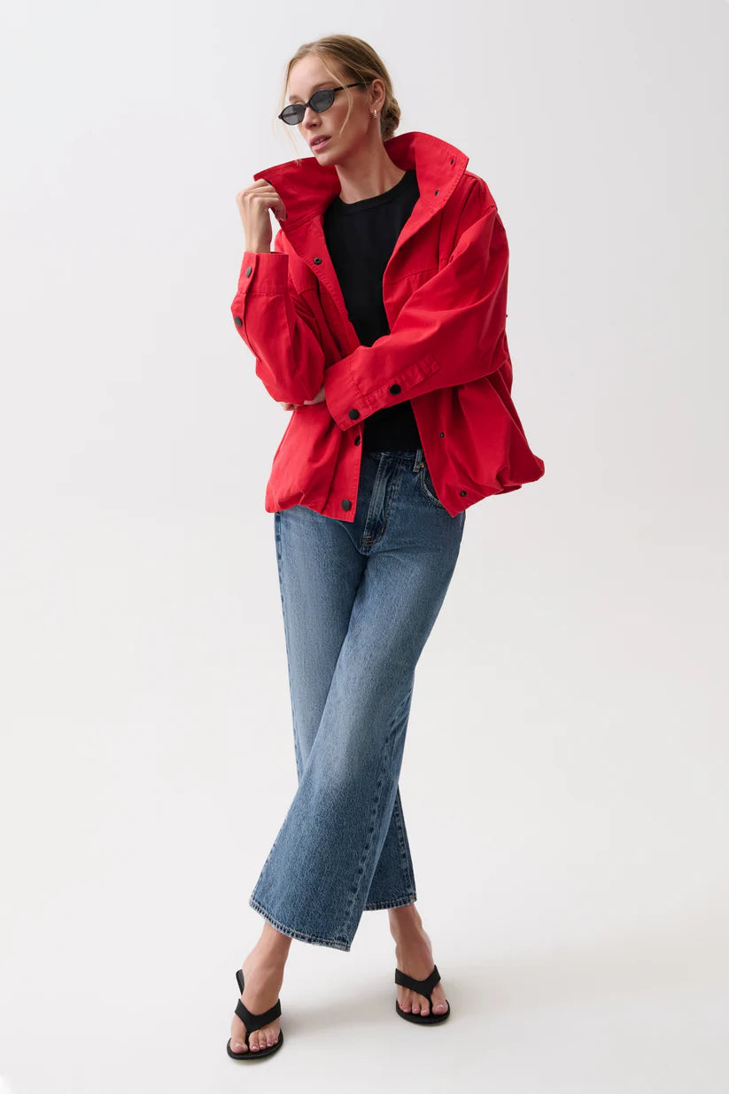 A woman modeling a bright red oversized jacket over a black top, paired with blue jeans and black sandals, against a plain white studio background.