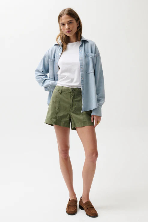 Woman wearing a light blue shirt, white top, green shorts, and brown shoes on a white background