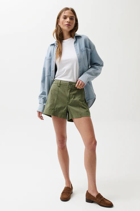 Woman wearing a light blue denim shirt, white t-shirt, green shorts, and brown loafers on a white background