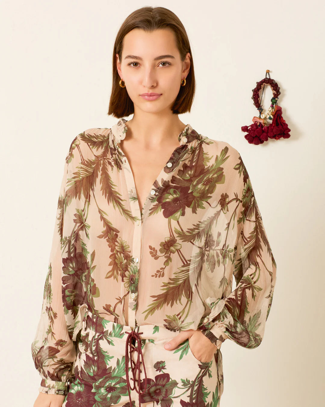 Woman wearing a floral blouse with a matching skirt, standing next to a red floral bag on a beige background.