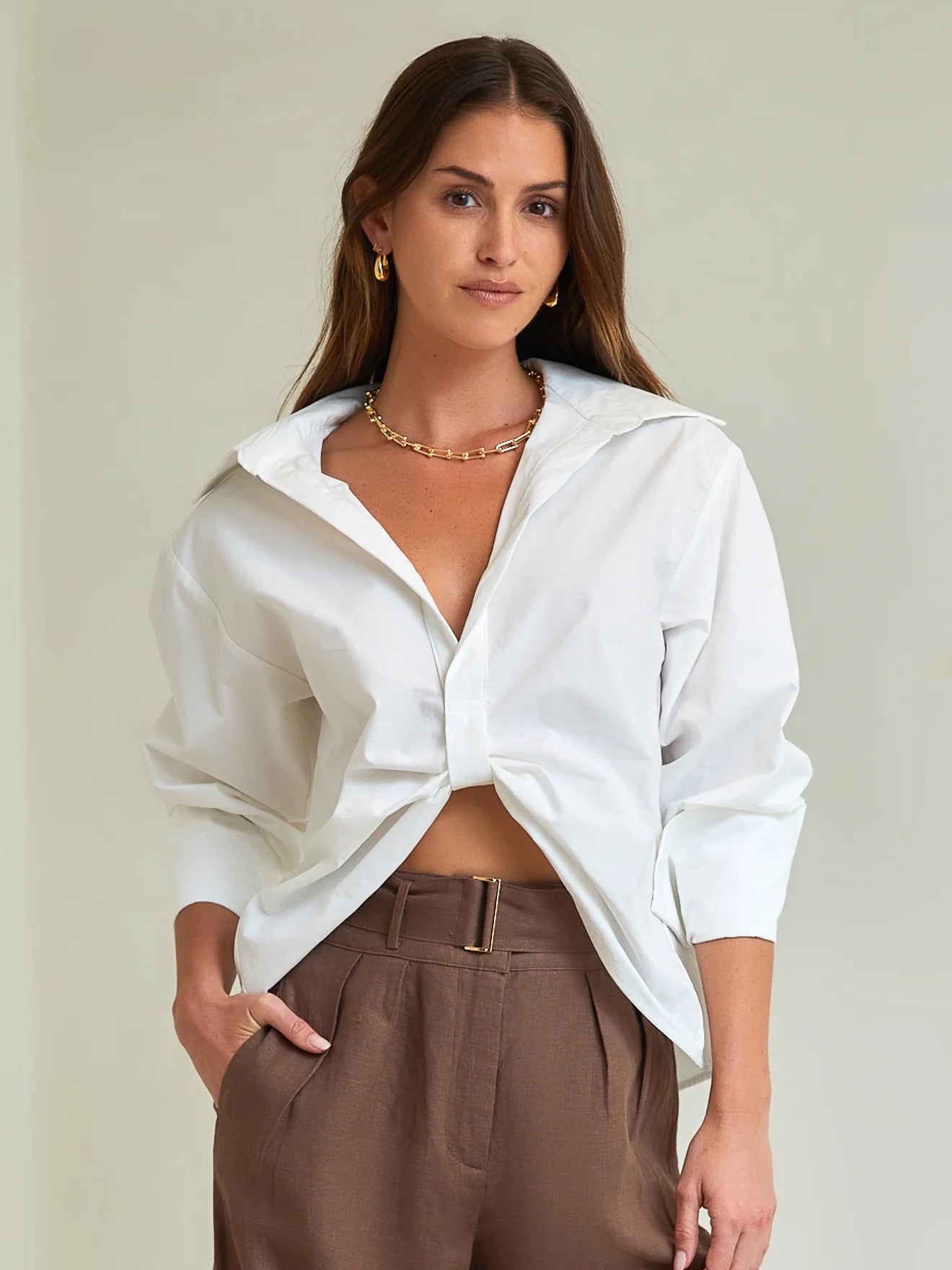 Woman wearing a white blouse with a knot detail and brown pants against a plain background