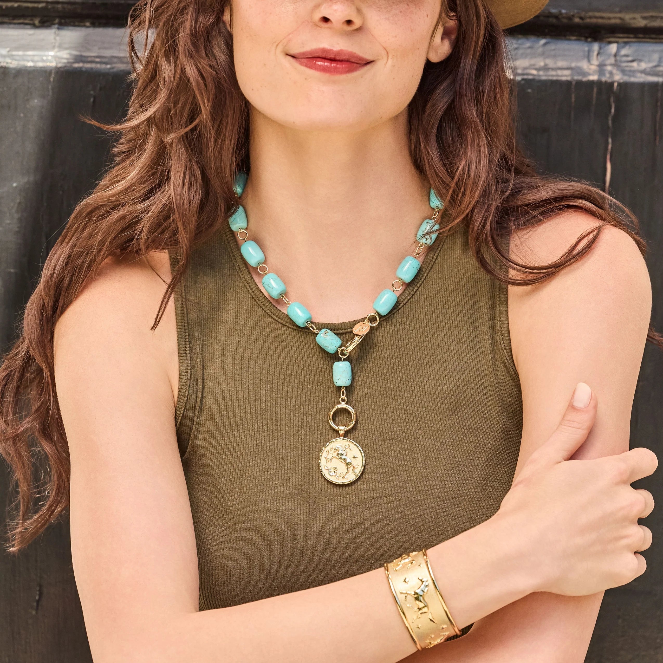 A woman wearing a turquoise and gold beaded necklace with a round coin pendant, styled with an olive green ribbed tank top and a gold cuff bracelet.