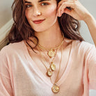 A close-up lifestyle shot of a woman wearing layered gold necklaces with circular sun and wave pendants over a light pink knit top.
