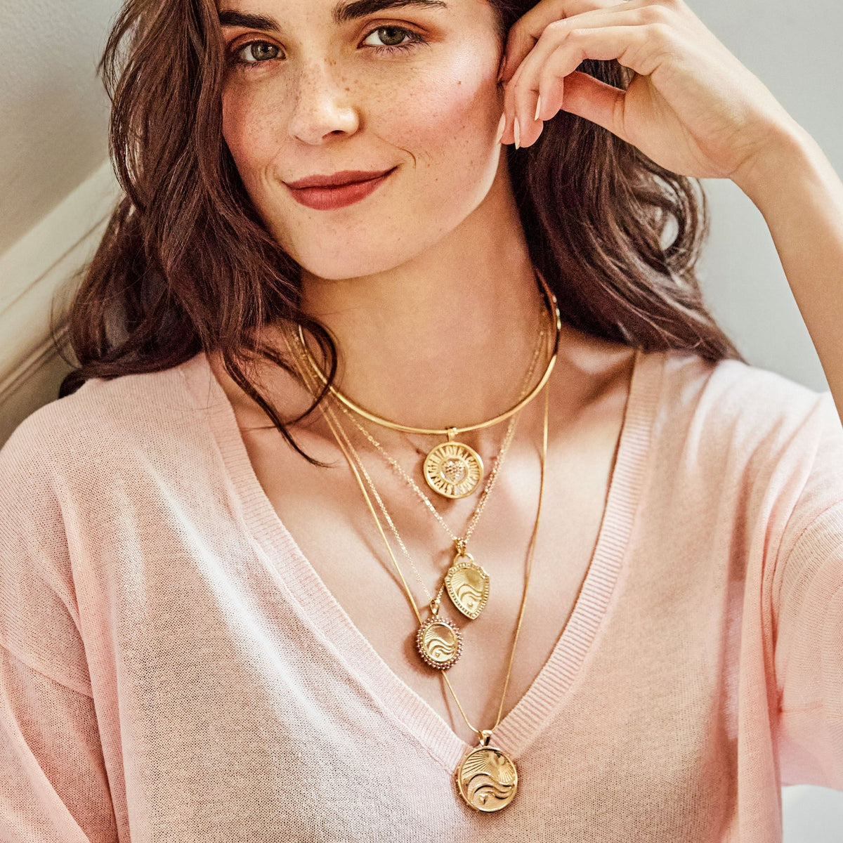 A close-up lifestyle shot of a woman wearing layered gold necklaces with circular sun and wave pendants over a light pink knit top.