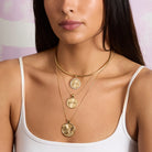 Woman wearing gold necklaces with a white top against a pink background