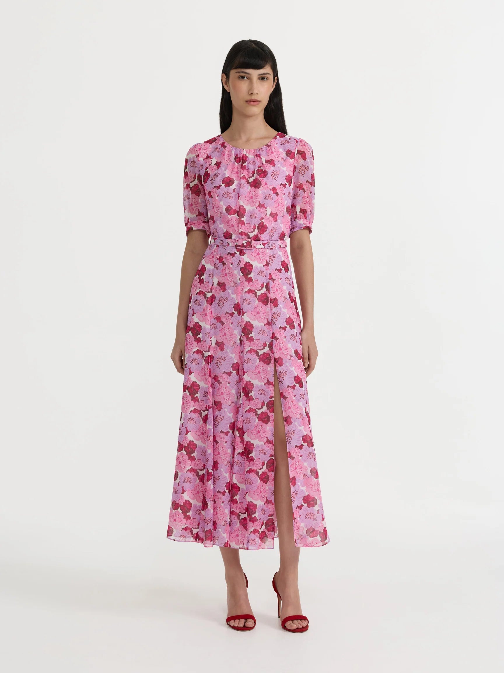 A model wearing a pink short-sleeved midi dress with a red and white floral pattern, standing against a plain white studio background.