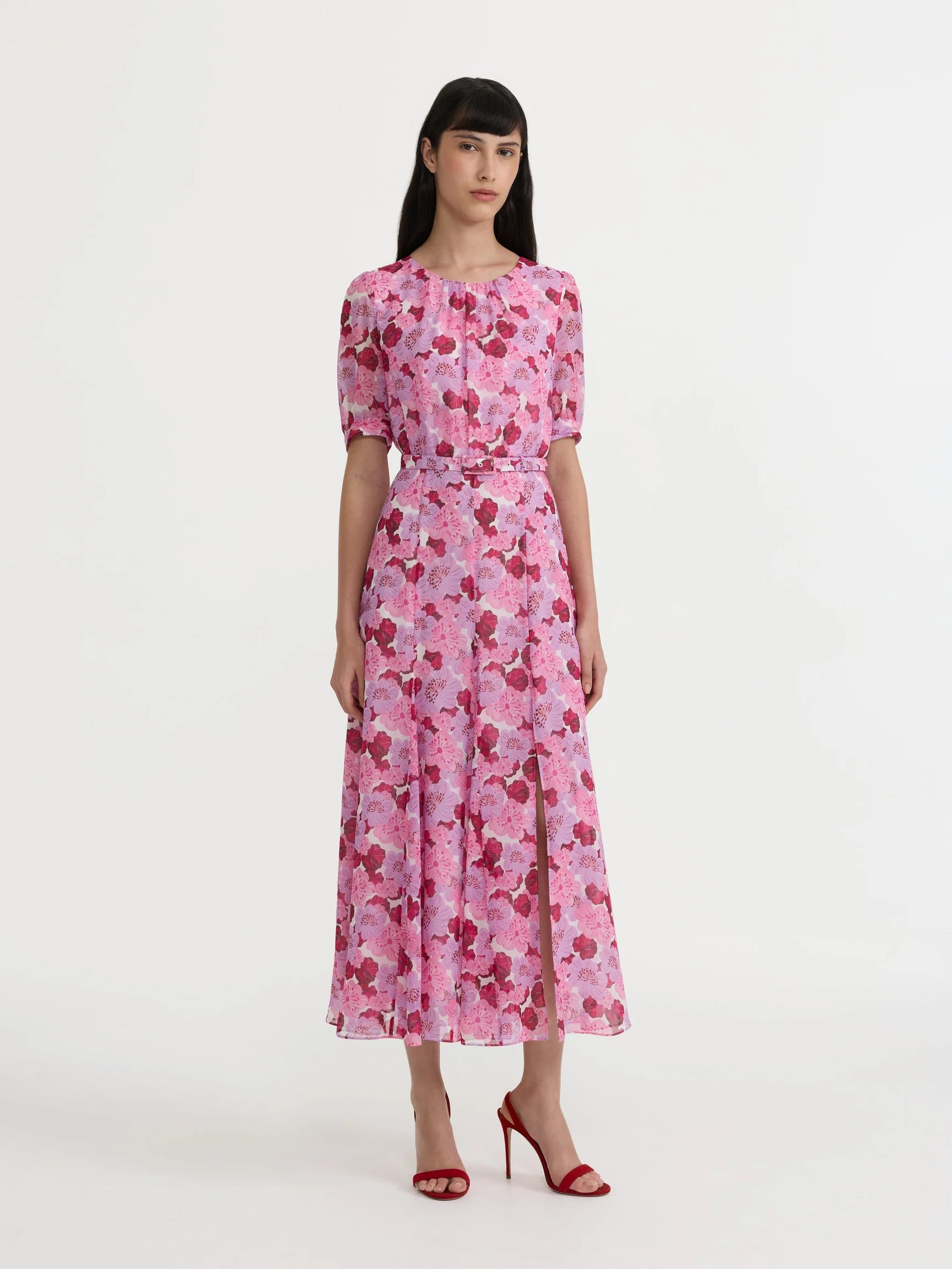 A woman modeling a pink floral midi dress with short puff sleeves, a defined waist, and a flared skirt, paired with red high-heeled sandals against a plain white studio background.