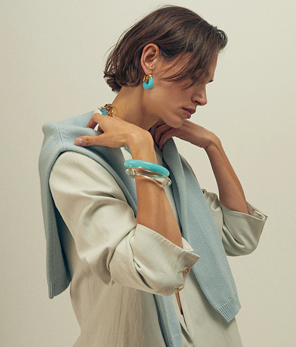 Woman wearing a light blue jacket and beige outfit with turquoise accessories against a neutral background