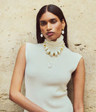 Woman wearing a white turtleneck and multiple necklaces against a beige stone wall.