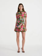 A model stands against a light gray studio background wearing a short-sleeve mini dress with a dense multicolor floral print and black strappy high-heeled sandals.