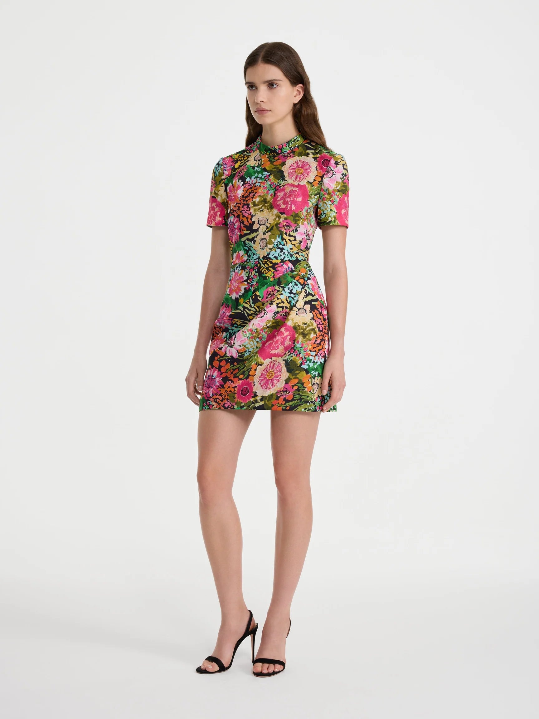 A model wearing a short-sleeved mini dress with a vibrant pink, red, and green floral print, paired with black high-heeled sandals, set against a plain light gray studio background.