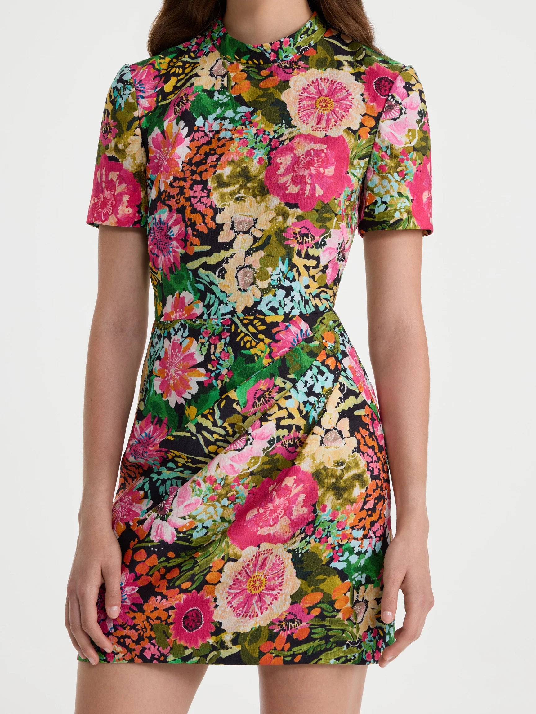 A woman wearing a short-sleeve, high-neck mini dress with a vibrant, dense floral print in pink, green, orange, and yellow on a dark background, photographed against a light gray studio backdrop.