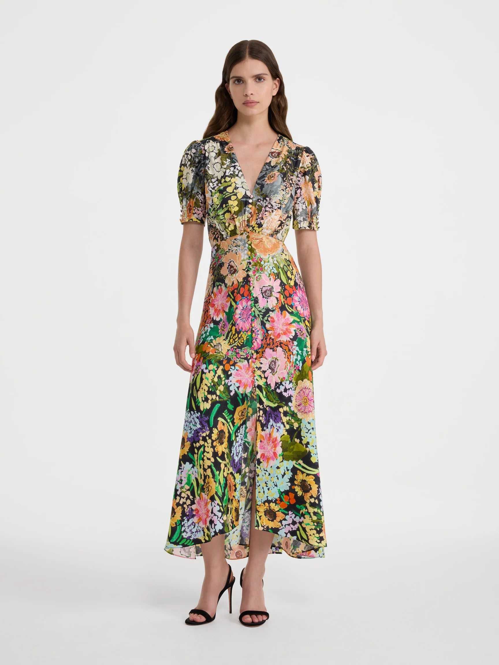 A model wearing a short-sleeved midi dress with a vibrant multicolor floral print, standing against a plain light gray studio background.